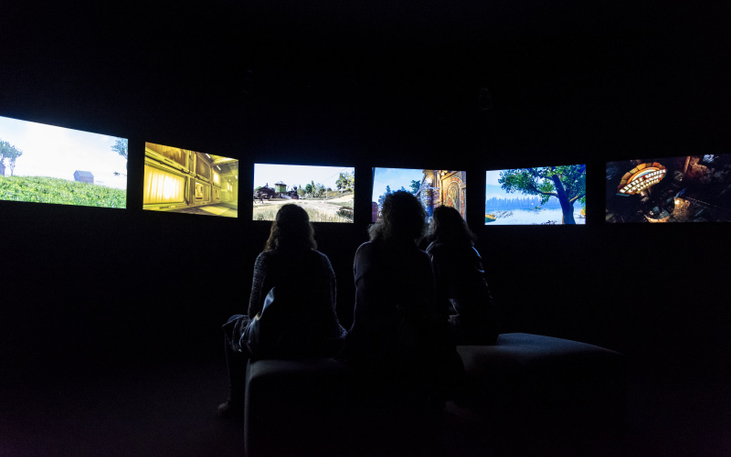 The photo shows three women sitting in a dark room, with only their silhouette visible. In a semicircle there is a row of screens surrounding the women.