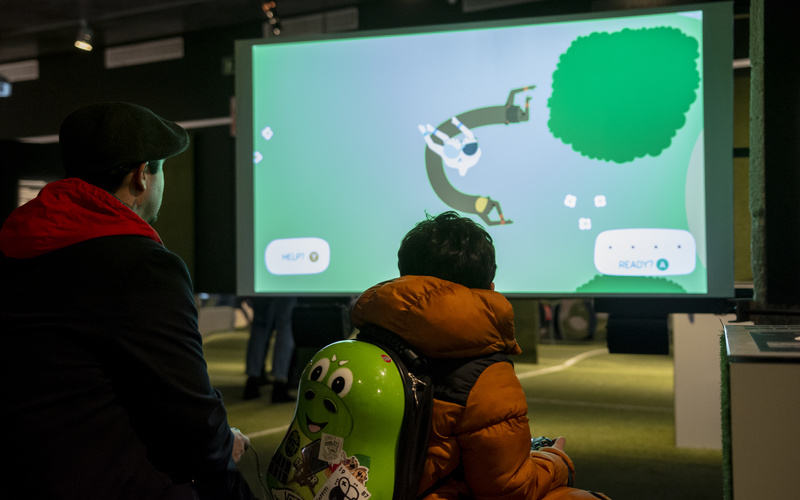 A man and a small boy are turned with their backs to the viewer. The boy is wearing a pear-shaped backpack, which embodies a computer game character. At the back of the picture there is a screen with a game, which is played. 