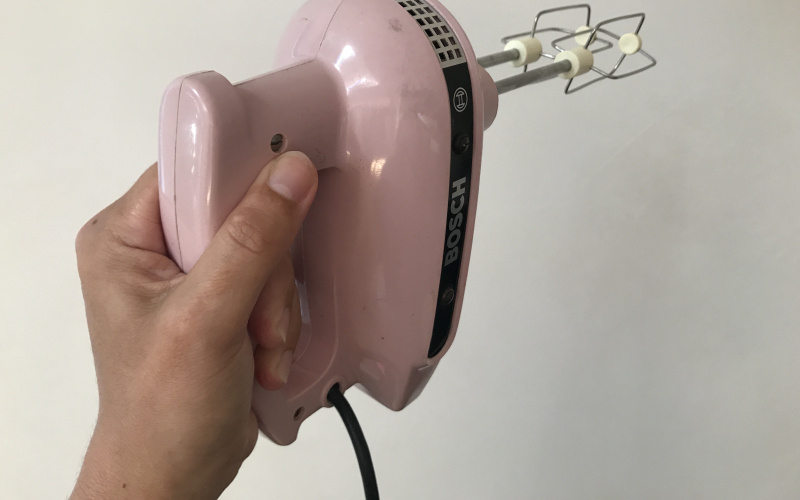 A pink hand mixer is lifted horizontally in front of a white wall.