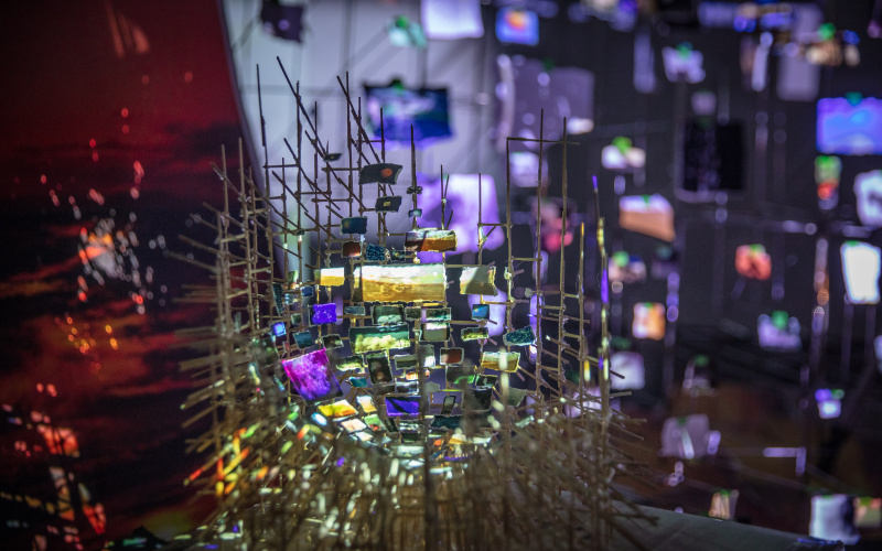 On display is a filigree installation of thin rods to which small pieces of paper are attached. On each piece of paper a light projection can be seen.