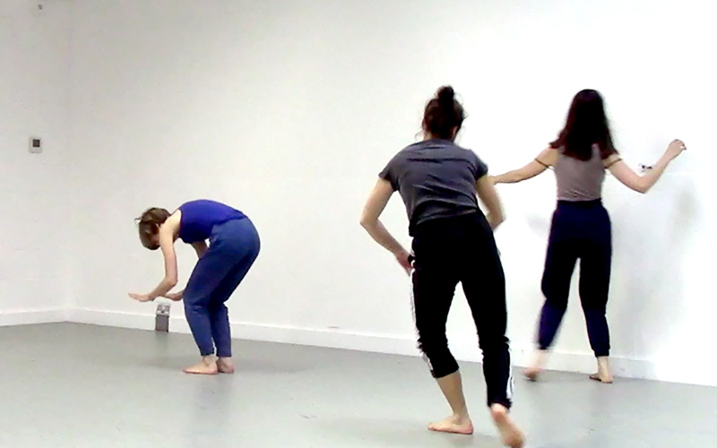 Zu sehen sind drei junge Frauen, die in tänzerischer Bewegung sich durch den Raum bewegen.