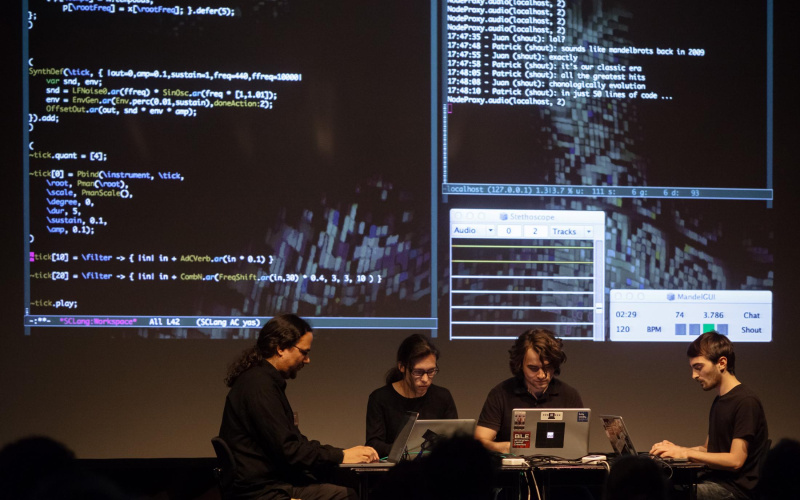 Four programmers sit in front of their computers while the produced data can be seen in the background on the screen