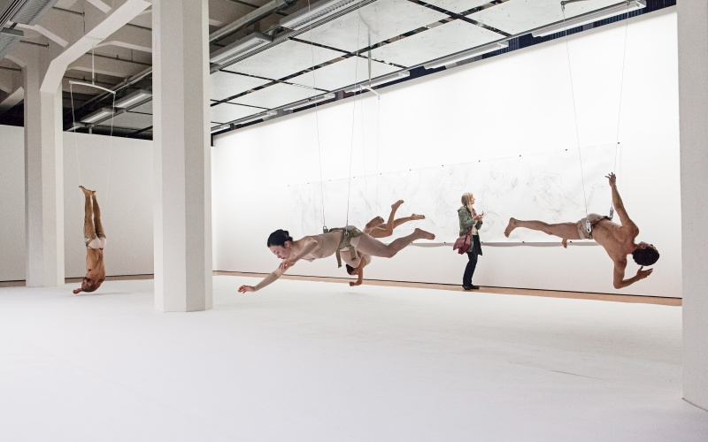 Five dancers hanging on ropes upside down in the exhibition space.