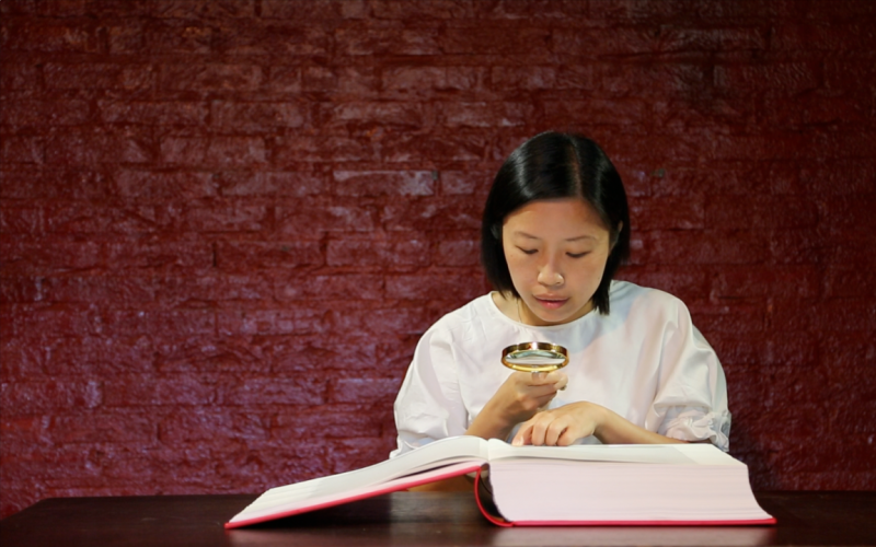 Eine Frau liest mit einer Lupe ein grosses aufgeschlagenes Buch vor einer roten Wand