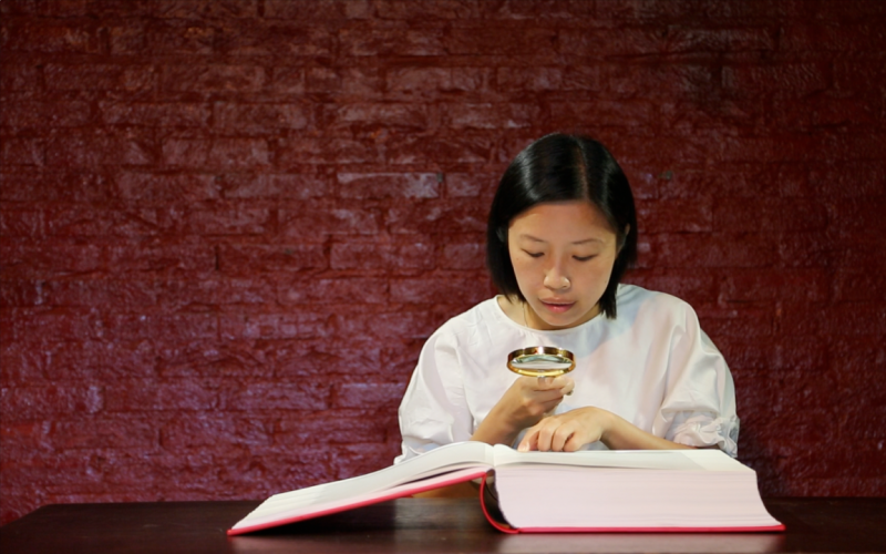 A woman reads a large open book in front of a red wall with a magnifying glass