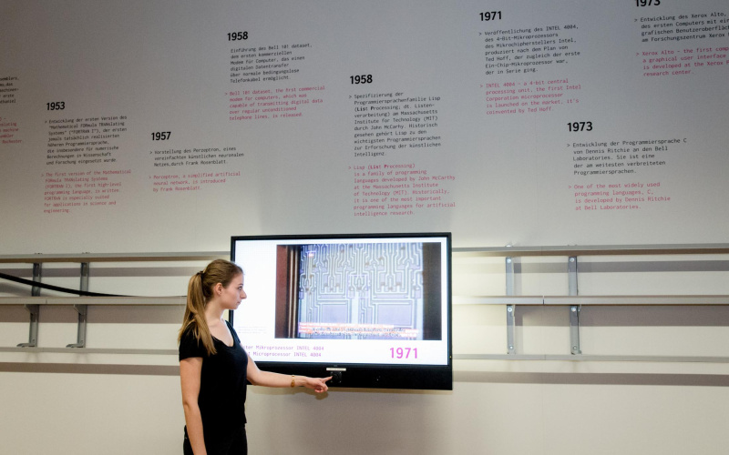 Das Bild zeigt eine Frau im Halbprofil, die die virtuelle Zeitleiste der »Genalogie des digitalen Codes« bedient. An der Wand, wo der Bildschirm auf einer beweglichen Schiene befestigt ist, ist eine Infografik zu sehen