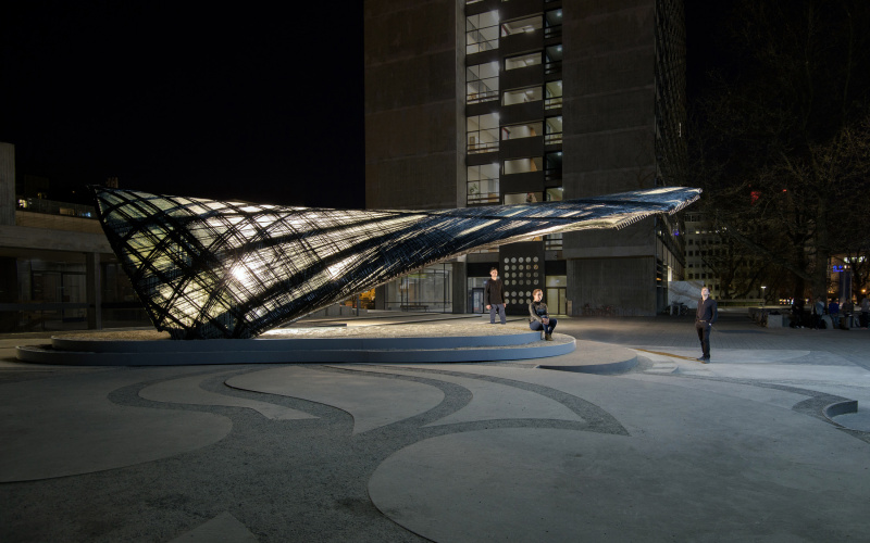 The picture shows a seated woman and two men standing around an illuminated research pavilion made of glass and carbon fibres