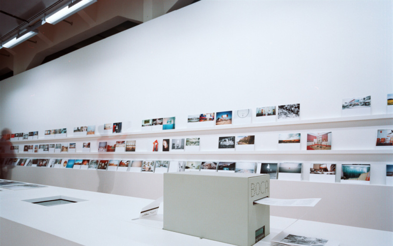 The picture shows a shelf with three levels where photographs are displayed. In the foreground you can see a photo printing machine