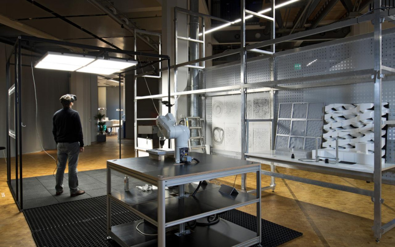 Installation with a robot and milled elements in grey shelves, next to which stands a man with VR glasses