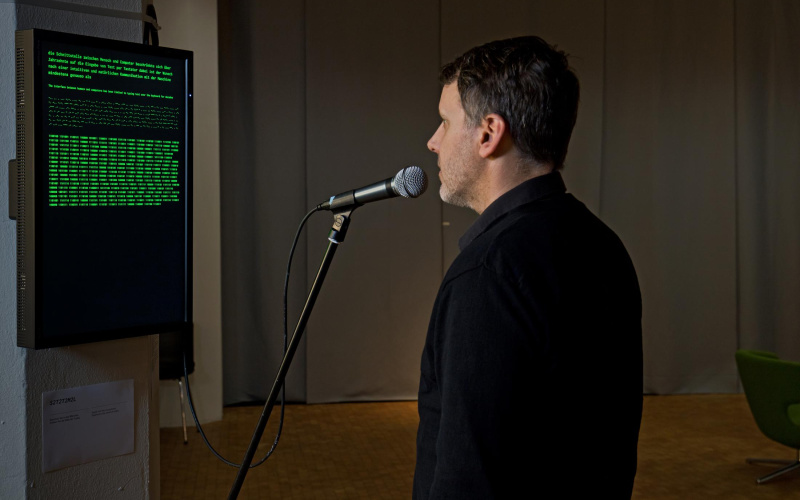 A man speaking into the microphone is standing in front of a screen with a German text.  Morse code and computer language can be seen
