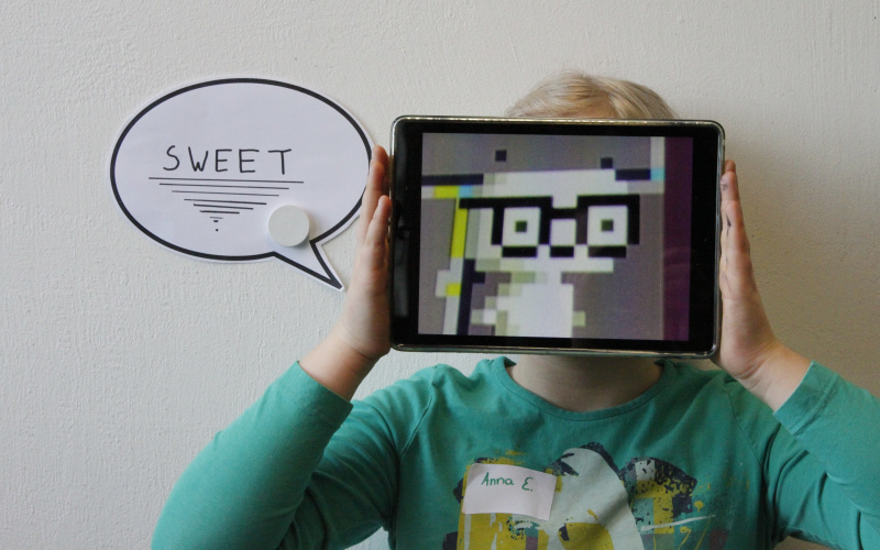 Performance of a girl, that is holding an iPad in front of her face. The display shows a creature buid of pixels. Next to her, the word "sweet" is written in a speech bubble.