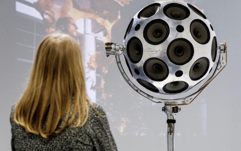 A woman stands in front of a silver sphere of loudspeakers