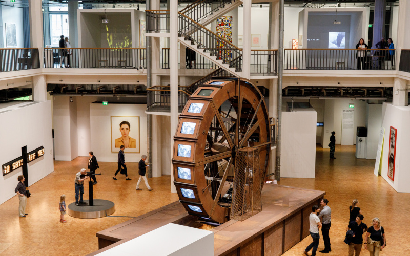 A huge metal mill wheel stands in an exhibition room. At the mill wheel there are tube screens on which videos of water are played.