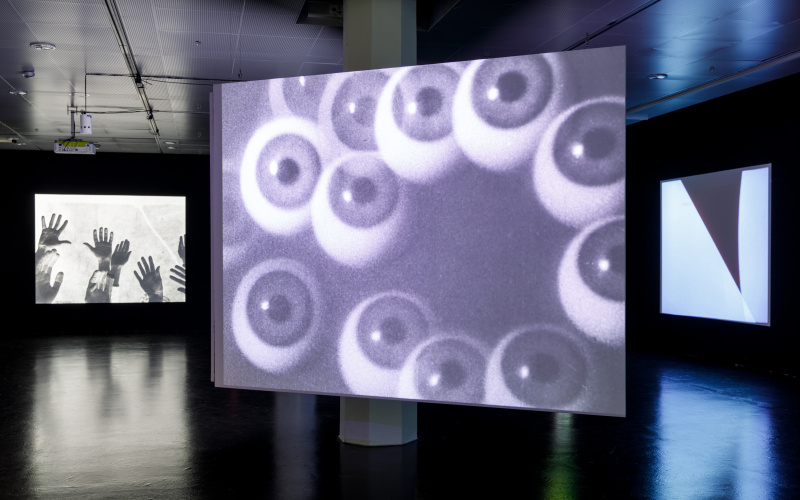 Das Foto zeigt den Ausstellungsraum mit einer großen Leinwand in der Mitte, die Kugeln abbildet, die wie Augäpfel aussehen. Links und rechts sind im Hintergrund zwei weitere Leinwände zu sehen, auf denen Hände und schwarz-weiße Formen abgebildet sind. 
