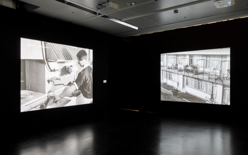 The photo shows the exhibition room with two large screens left and right on the walls. The left canvas shows a woman doing the dishes, the right canvas shows a retirement home from outside and people sitting on balconies. 