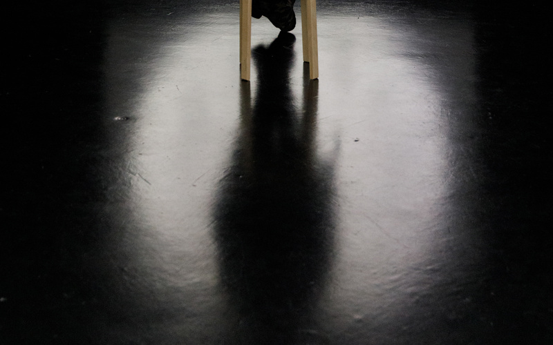 The photo shows the silhouette of a person watching a black and white film on a large screen. On the screen, an analogue clock strikes twelve. The light of the screen falls a round blurred light on the floor.