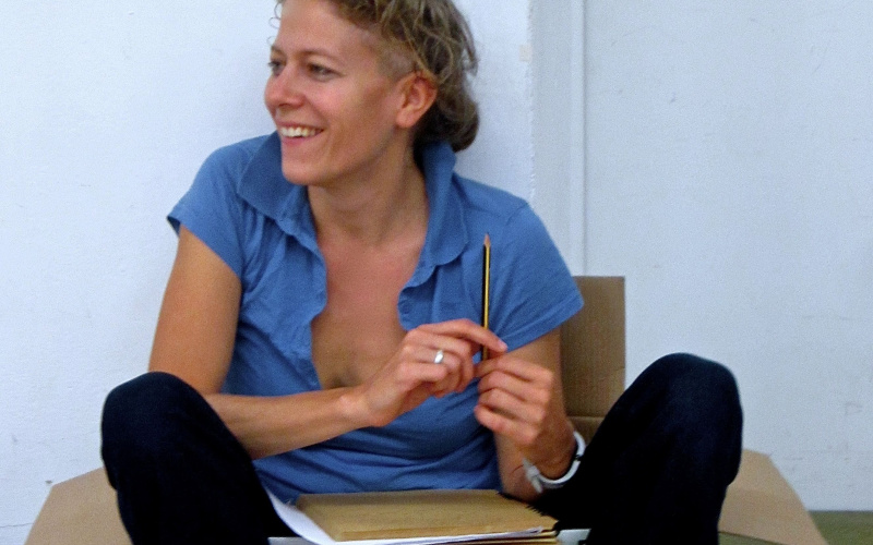 Anja Konjetzky sitzt lässig in einem Pappkarton und hält einen Bleistift in der Hand. 
