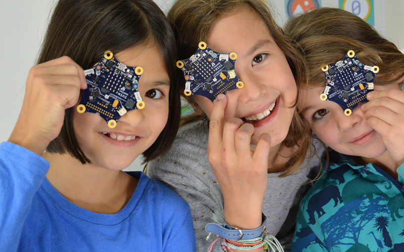 Photo of three young girls each holding a Calliope mini circuit board in front of their face.