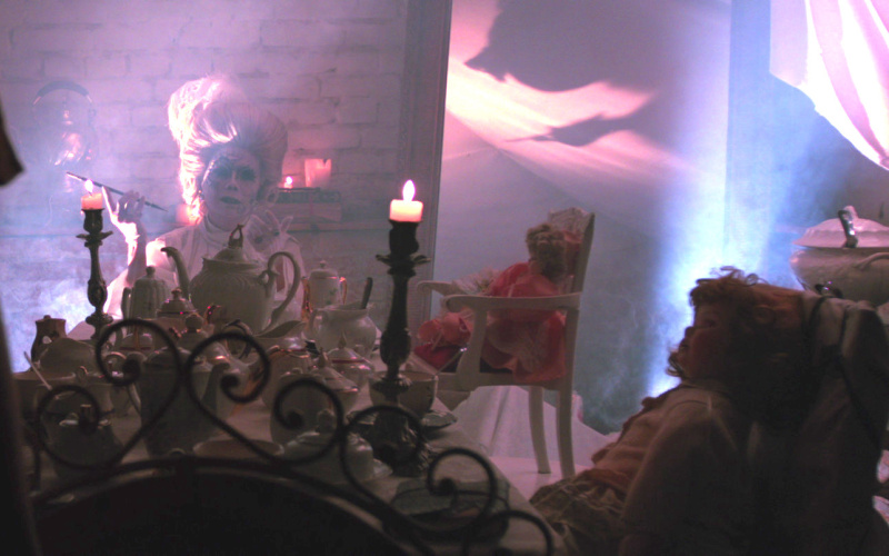 Eine rauchende Person sitzt in einem Raum mit Steinwänden. Sie hat eine Perücke aus dem 18. Jahrhundert auf, ist stark geschminkt und sitzt an einem Tisch auf dem eine lange Kerze brennt. Um den Tisch sitzen Puppen.