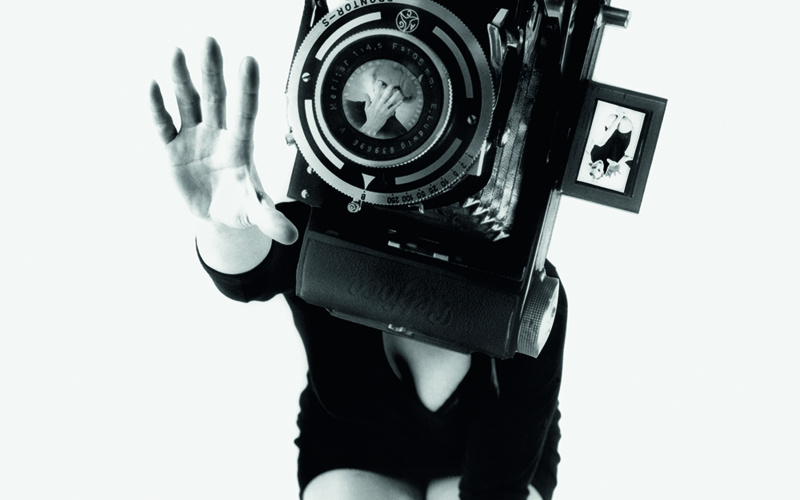 Das Schwarzweiß-Photo zeigt eine Frau in einem schwarzen Minikleid auf den Knien. Ihr Gesicht ist von einer überdimensional großen Kamera verdeckt. 