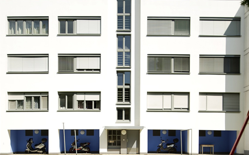 Zu sehen ist ein großes Weiße Gebäude mit einer Galerie im Erdgeschoss, deren Wände blau gestrichen sind. Das Gebäude befindet sich in der Dammerstocksiedlung in Karlsruhe, die aus der Bauhaus Zeit stammt.