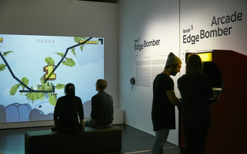 Exhibition view »EdgeBomber« with players in front of a wall projection and a game machine