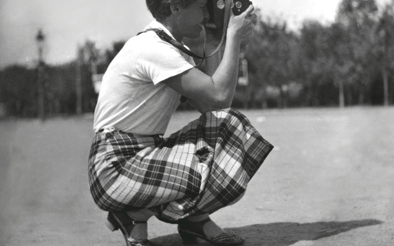 The black and white photograph shows a woman in squatting position with a camera.