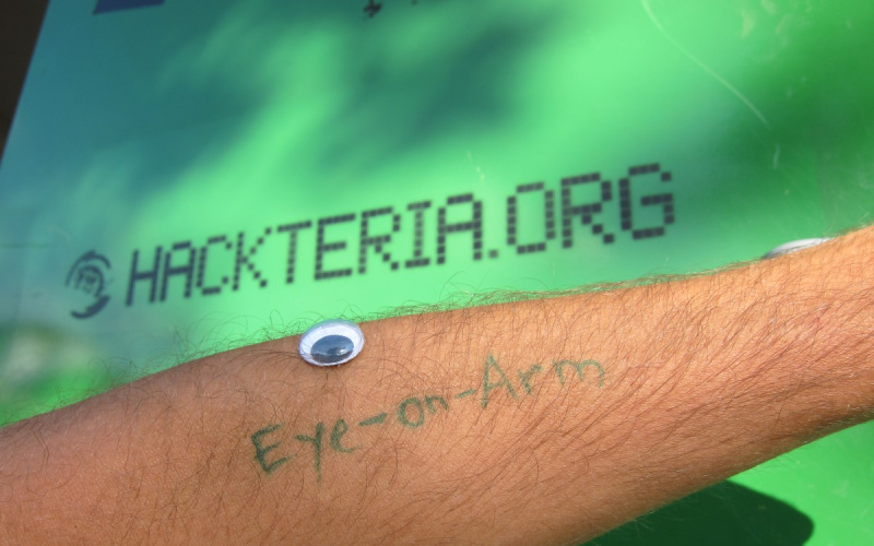 Auf einem Männerarm ist mit grüner Schrift Eye-on-Arm geschrieben und ein Spielzeugauge liegt darüber auf dem Unterarm. Der Hintergrund ist grün und mit der url hackteria.org beschrieben.