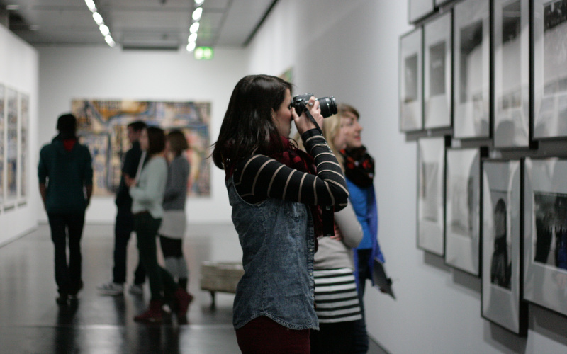 Eine Frau steht vor mehreren Bildern, die im Museum an der Wand hängen. Sie hält ihre Kamera vor das Gesicht und fotografiert die Fotografien.