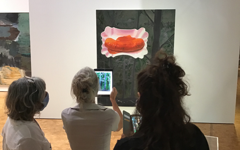 Three women stand in front of a picture showing a curry sausage in a cardboard bowl. One of the women holds an iPad on the picture.