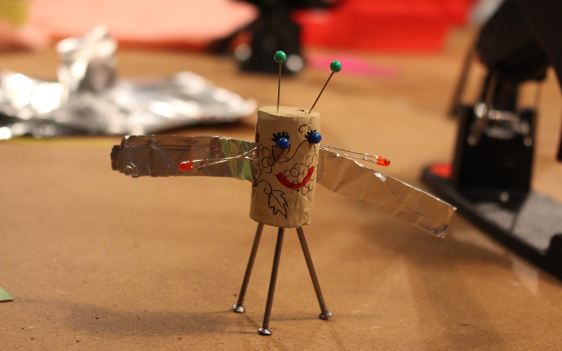 A kork, that has been turned into a little figurine with wings is sitting on a table with crafting materials around it.