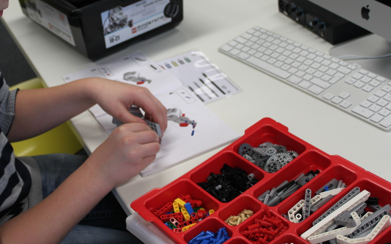 A boy is constructing a lego-robot. On the table he is sitting at are boxes filled with robot parts.