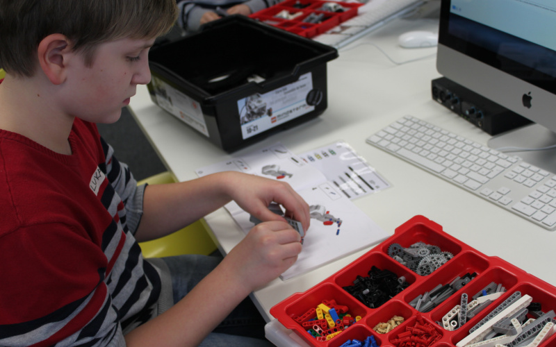 A boy is constructing a lego-robot. On the table he is sitting at are boxes filled with robot parts.