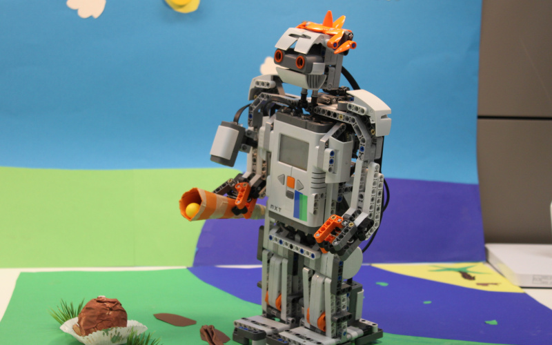 Vor einer bunten Papierkulisse, die Rasen eine Sonne und Wolken andeutet, steht ein Legoroboter.