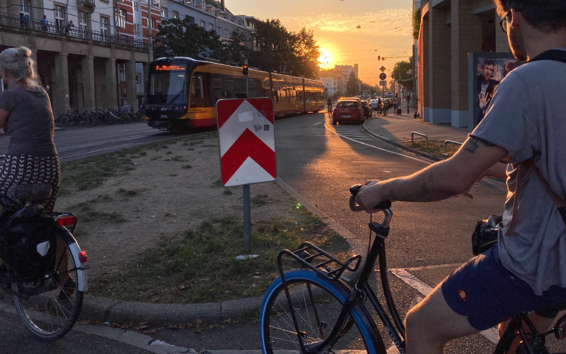 In the foreground, a young man on a bicycle is riding along a road. In front of him, the setting sun is shining and illuminating the streetcar that crosses the landscape.