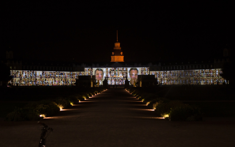  The facade of Karlsruhe´s Castle is projected with many passport photographs of various people.
