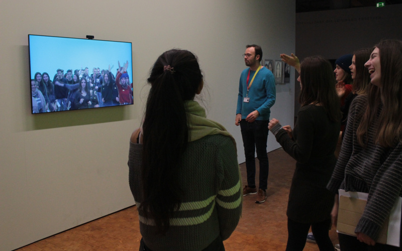 A group of young people can be seen standing in front of a screen. The screen shows a live image of the group, which is recorded by a camera above the screen.
