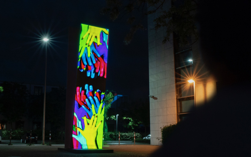 Many hands are projected on a column. One set of hands stretches from bottom to top towards the other set of hands and vice versa. It is night.