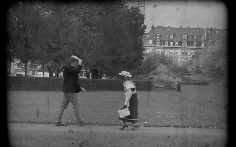 Zwei Passanten begegnen sich auf der Straße, der Herr lüpft den Hut um die sich nähernde Dame zu grüßen. Es ist ein Standbild aus einem schwarzweiß Film, der bei dem Workshop "Die kleinen Strolche" entstanden ist.