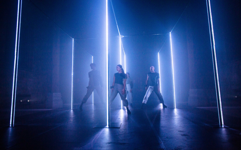A group of dancers stands in a darkened room illuminated by blue neon tubes.