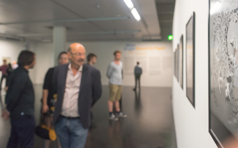People in the museum at the Vernissage are blurry in the picture, but the artwork on the wall is sharp and shows abstract forms in shades of gray.