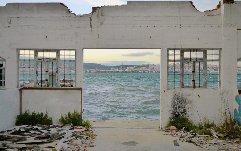 Das Bild zeigt eine Hausruine, durch deren offene Türen und Fenster man auf das Meer und eine dahinter liegende Stadt sehen kann. 