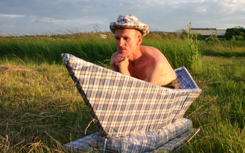 Ein Mann mit nacktem Oberkörper sitz in einem überdimensional großen Papierboot, das auf einer Wiese steht