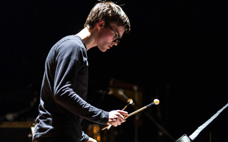  Young man with glasses and dark long-sleeved shirt plays on percussion.