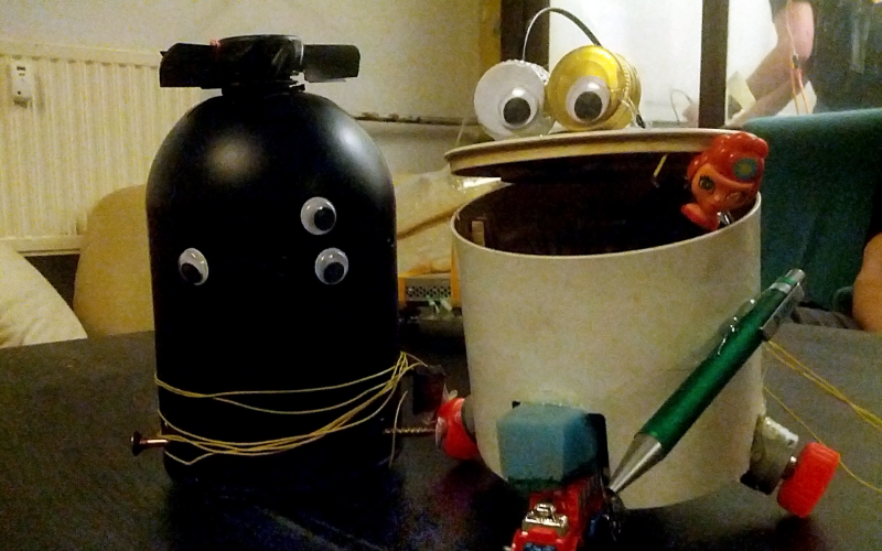 Two little robots that are made out of junk are sitting next to each other, one is black and has 3 eyes and one is made out of a bin with 2 eyes out of bottle caps, that are placed on the open mouth resp. lid of the bucket.