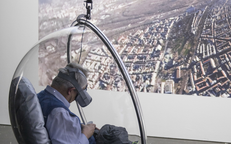 A visitor with VR glasses sits in a hanging semicircular armchair made of transparent plastic.