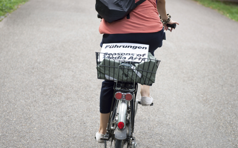 In einem Fahrradkorb liegt ein weißes, laminiertes Schild, darauf steht »Führungen Seasons of Media Arts«