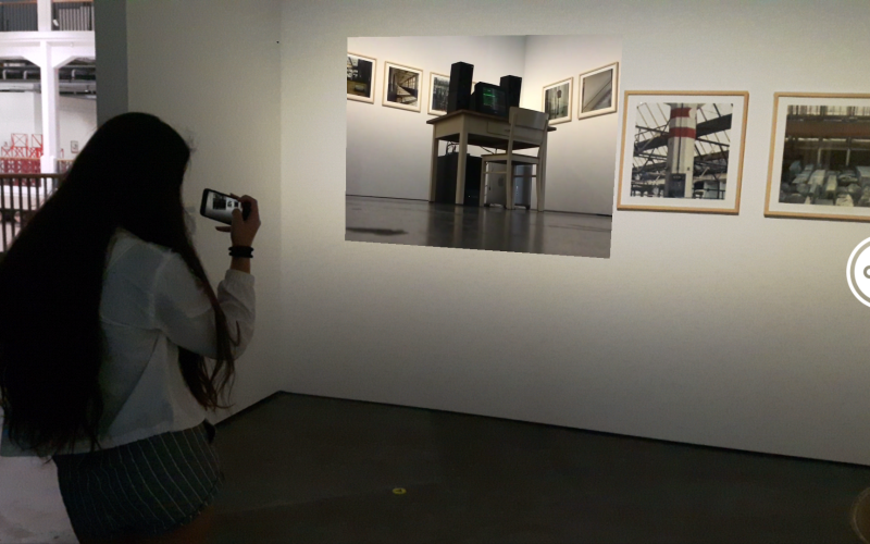 Ein Mädchen steht mit ihrem Handy in der Hand vor einem Bild. Sie hält ihr Handy so als würde sie das Bild filmen.