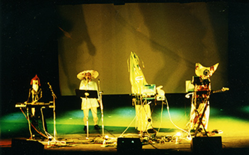 Four figures standing on a stage with colorful costumes and instruments.