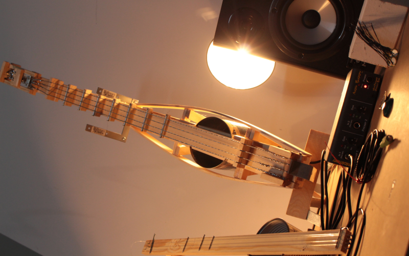 A diy guitar out of thin wood and a tin can is standing upright next to a speaker. A bright light is shining on the instrument from behind.
